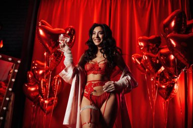 Sensual curly black haired woman in fishnet gloves with glass of wine posing on red glamour background 