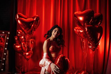 Young tempting brunette woman with perfect skin in sexy lingerie posing with heart balloons in a red room  