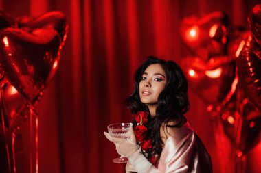 Sensual curly black haired woman in fishnet gloves with glass of wine posing on red glamour background 