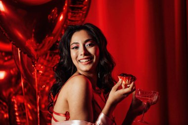 Sensual young japanese black hair woman in sexy lingerie posing with cupcake and coupe glass of champagne at red glamour background with balloons