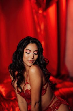 Side view of curly black haired asian sensual model seduces sitting on the kneel on the floor at red glamour background