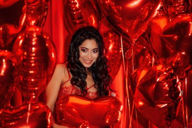 Young tempting brunette woman with perfect skin in sexy lingerie posing with heart balloons in a red room  