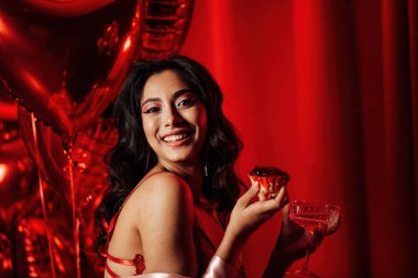 Sensual young japanese black hair woman in sexy lingerie posing with cupcake and coupe glass of champagne at red glamour background with balloons