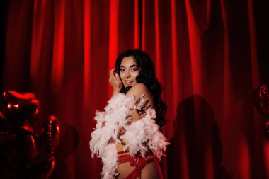 Sensual curly black hair asian woman with feather boa posing on red glamour background