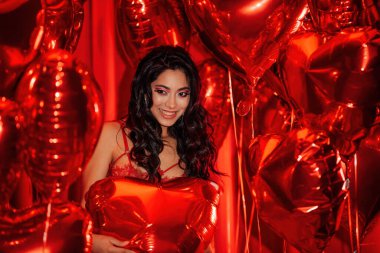 Attractive tempting brunette woman with perfect skin in sexy lingerie posing with heart balloons in a red room  