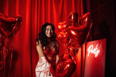 Sensual curly black hair woman with perfect skin in sexy lingerie posing with heart balloons in a red room  