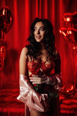 Sensual asian woman with perfect skin in sexy lingerie posing with heart balloons in a red room  