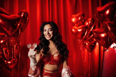 Curly black haired woman in fishnet gloves and lace underwear with glass of wine posing on red glamour background 