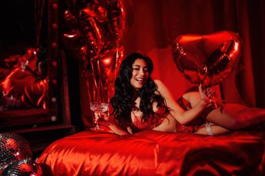 Beautiful japanese lady in red underwear lying on silk sheet with champagne glass and balloon heart, posing on Valentine's day