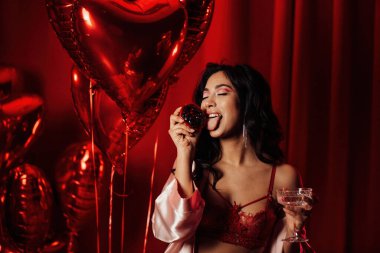 Sensual young japanese black hair woman in sexy lingerie posing with cupcake and coupe glass of champagne at red glamour background with balloons