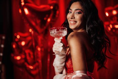 Sensual asian black haired model in lace underwear and fishnet gloves posing with coupe glass of champagne on red glam background.