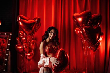 Young tempting brunette woman with perfect skin in sexy lingerie posing with heart balloons in a red room  