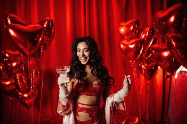 Sensual curly black haired woman in fishnet gloves with glass of wine posing on red glamour background 