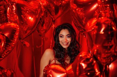 Smiling young tempting brunette woman with perfect skin in sexy lingerie posing with heart balloons in a red room  