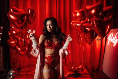 Sensual asian black haired model in lace underwear and fishnet gloves posing with coupe glass of champagne on red glam background.