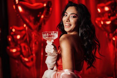 Sensual asian black haired model in lace underwear and fishnet gloves posing with coupe glass of champagne on red glam background.