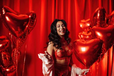 Happy young tempting brunette woman with perfect skin in sexy lingerie posing with heart balloons in a red room  
