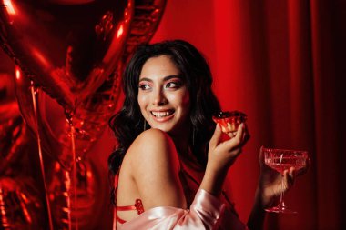 Sensual young japanese black hair woman in sexy lingerie posing with cupcake and coupe glass of champagne at red glamour background with balloons