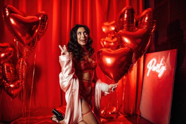 Happy brunette woman in lace underwear posing with heart balloons in a red room   
