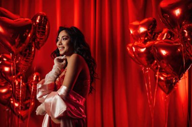 Tempting brunette woman with perfect skin in sexy lingerie posing with heart balloons in a red room  