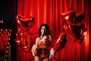 Young tempting brunette woman with perfect skin in sexy lingerie posing with heart balloons in a red room  