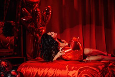 Portrait of sexy japanese lady in red underwear lying on silk sheet with balloon heart, posing on Valentine's day