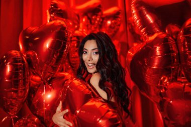 Smiling brunette japanese woman with perfect skin in sexy lingerie posing with heart balloons in a red room 