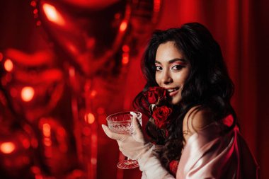 Brunette woman in sexy underwear posing with roses and coupe glass of champagne in a room decorated for Valentine's Day