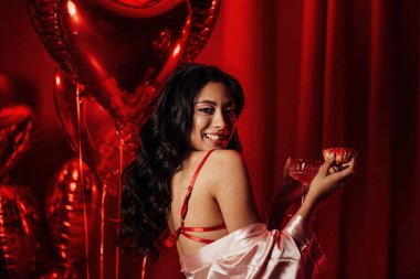 Sensual young japanese black hair woman in sexy lingerie posing with cupcake and coupe glass of champagne at red glamour background with balloons