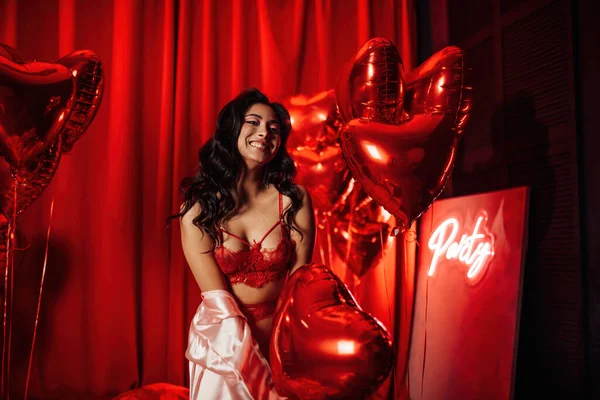 Joyful hot asian model with perfect skin in sexy lingerie posing with heart balloons in a red room