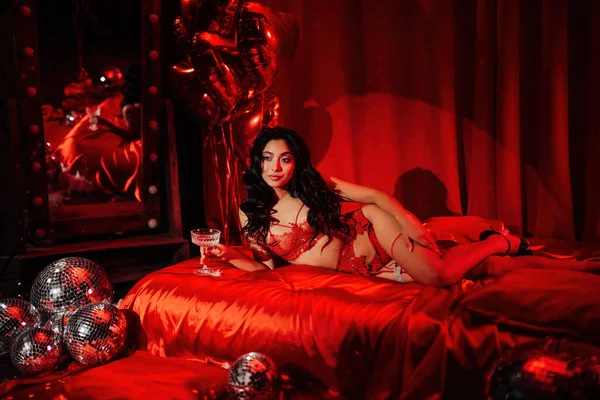 Sexy japanese lady in red underwear lying on silk sheet with champagne glass, posing on Valentine's day