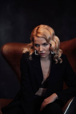 Young adult beauty woman in formal evening suit of black color with lace bra at thoughtful. Stylish blonde curly hair sensual model fashionista posing at studio in fashion pantsuit