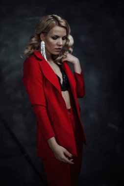 Young adult beauty woman in formal evening suit of red color with lace black bra underwear standing by thoughtful. Stylish blonde curly hair model fashionista posing at studio in fashion pantsuit