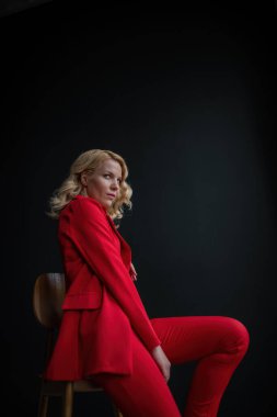 Young adult beauty woman in formal evening suit of red color with lace black bra underwear standing by thoughtful. Stylish blonde curly hair model fashionista posing at studio in fashion pantsuit