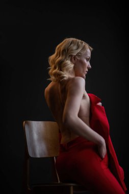 Adult beauty woman bare back in formal evening red trousers sitting pose without bra. Stylish blonde curly hair sensual nude model fashionista posing at studio in fashion pantsuit out of blazer