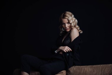 Young adult beauty woman in formal evening suit of black color with lace bra at thoughtful. Stylish blonde curly hair sensual model fashionista posing at studio in fashion pantsuit
