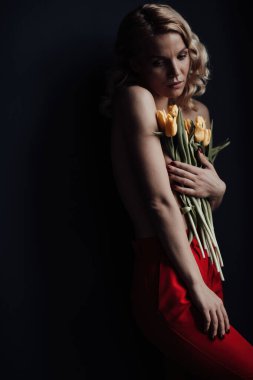 Adult beauty woman half naked in formal evening red trousers without bra hugs bouquet of yellow tulips. Stylish blonde curly hair sensual nude model fashionista posing in studio at spring holidays