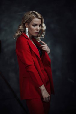 Young adult beauty woman in formal evening suit of red color with lace black bra underwear standing by thoughtful. Stylish blonde curly hair model fashionista posing at studio in fashion pantsuit
