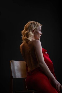 Adult beauty woman bare back in formal evening red trousers sitting pose without bra. Stylish blonde curly hair sensual nude model fashionista posing at studio in fashion pantsuit out of blazer