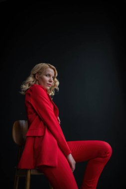 Young adult beauty woman in formal evening suit of red color with lace black bra underwear standing by thoughtful. Stylish blonde curly hair model fashionista posing at studio in fashion pantsuit