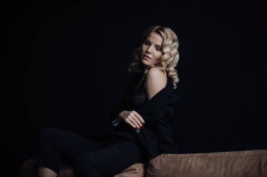 Young adult beauty woman in formal evening suit of black color with lace bra at thoughtful. Stylish blonde curly hair sensual model fashionista posing at studio in fashion pantsuit