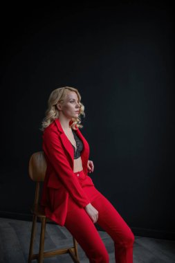 Young adult beauty woman in formal evening suit of red color with lace black bra underwear standing by thoughtful. Stylish blonde curly hair model fashionista posing at studio in fashion pantsuit