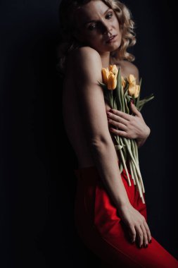 Adult beauty woman half naked in formal evening red trousers without bra hugs bouquet of yellow tulips. Stylish blonde curly hair sensual nude model fashionista posing in studio at spring holidays