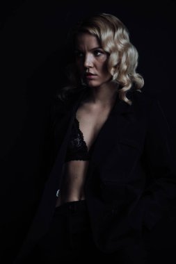 Young adult beauty woman in formal evening suit of black color with lace bra at thoughtful. Stylish blonde curly hair sensual model fashionista posing at studio in fashion pantsuit