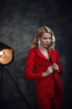 Young adult beauty woman in formal evening suit of red color with lace black bra underwear standing by thoughtful. Stylish blonde curly hair model fashionista posing at studio in fashion pantsuit