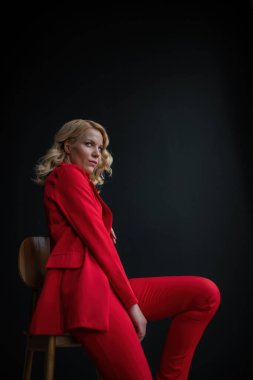 Young adult beauty woman in formal evening suit of red color with lace black bra underwear standing by thoughtful. Stylish blonde curly hair model fashionista posing at studio in fashion pantsuit