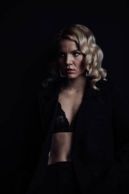 Young adult beauty woman in formal evening suit of black color with lace bra at thoughtful. Stylish blonde curly hair sensual model fashionista posing at studio in fashion pantsuit