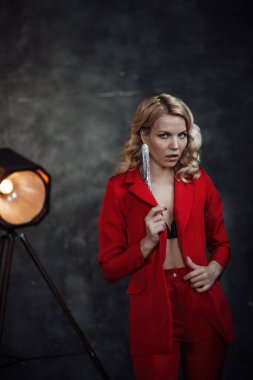 Young adult beauty woman in formal evening suit of red color with lace black bra underwear standing by thoughtful. Stylish blonde curly hair model fashionista posing at studio in fashion pantsuit