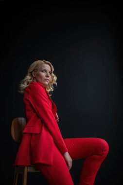 Young adult beauty woman in formal evening suit of red color with lace black bra underwear standing by thoughtful. Stylish blonde curly hair model fashionista posing at studio in fashion pantsuit