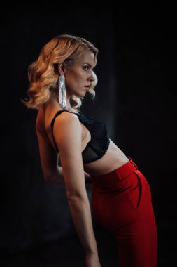 Young adult beauty woman in formal evening suit of red color with lace black bra underwear standing by thoughtful. Stylish blonde curly hair model fashionista posing at studio in fashion pantsuit
